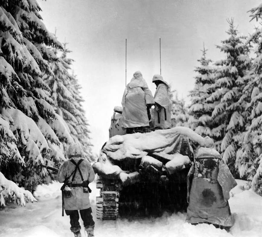 Troops advancing behind a Sherman tank during the Ardennes Offensive.